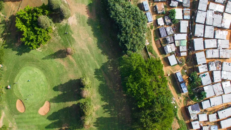 Eine Luftaufnahme in Vogelperspektive („Top-down“), die eine scharfe Grenze zwischen einem üppigen, grünen Golfplatz mit Sandbunkern auf der linken Seite und einer dicht gedrängten informellen Siedlung (Slum) mit Wellblechdächern auf der rechten Seite zeigt. Foto von Johnny Miller