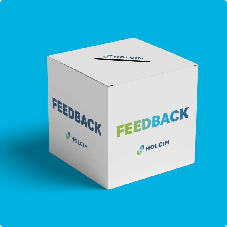 Eine weiße, würfelförmige Feedback-Box mit einem Schlitz an der Oberseite. Das Wort ‚FEEDBACK‘ ist in großen Buchstaben mit einem Blau-Grün-Verlauf auf die Seiten gedruckt, begleitet vom Holcim-Logo.
