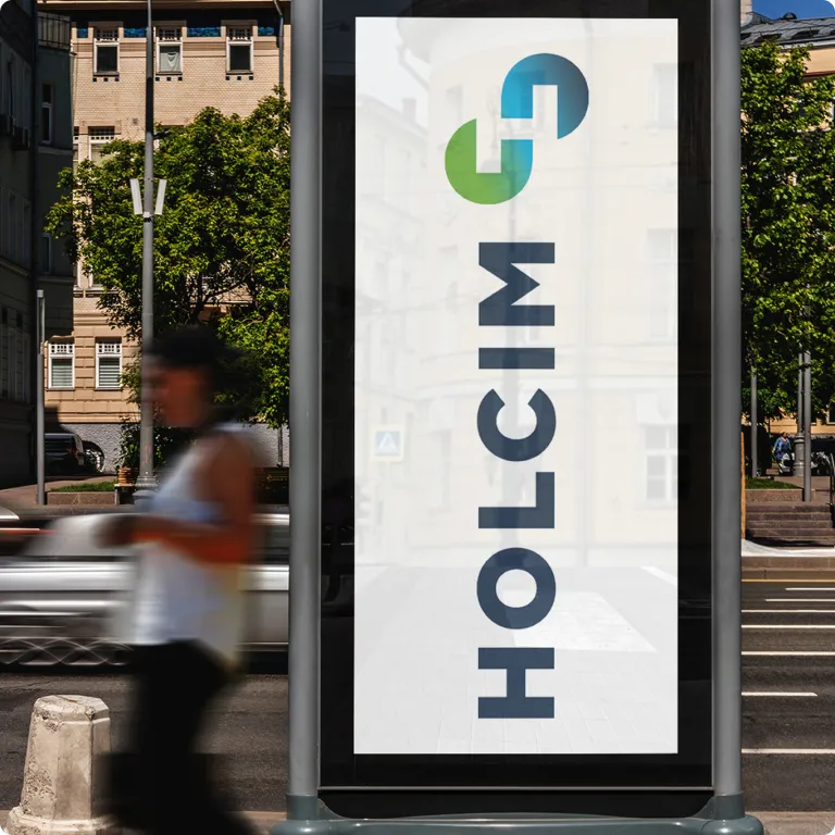 Ein hohes, vertikales Straßenbanner in einer städtischen Umgebung. Das Banner ist weiß und zeigt das Holcim-Logo sowie das Unendlichkeitssymbol vertikal in Dunkelblau und einem Grün-Blau-Verlauf.