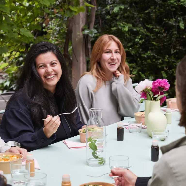 Arbeitskolleginnen lachen und essen zusammen an einem Tisch im Freien.