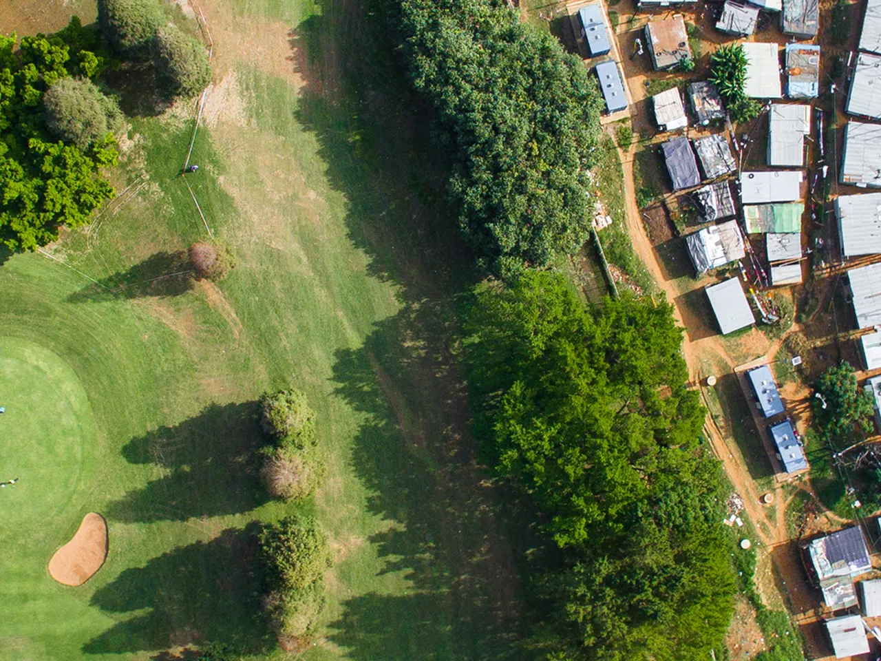 Eine Luftaufnahme in Vogelperspektive („Top-down“), die eine scharfe Grenze zwischen einem üppigen, grünen Golfplatz mit Sandbunkern auf der linken Seite und einer dicht gedrängten informellen Siedlung (Slum) mit Wellblechdächern auf der rechten Seite zeigt. Foto von Johnny Miller