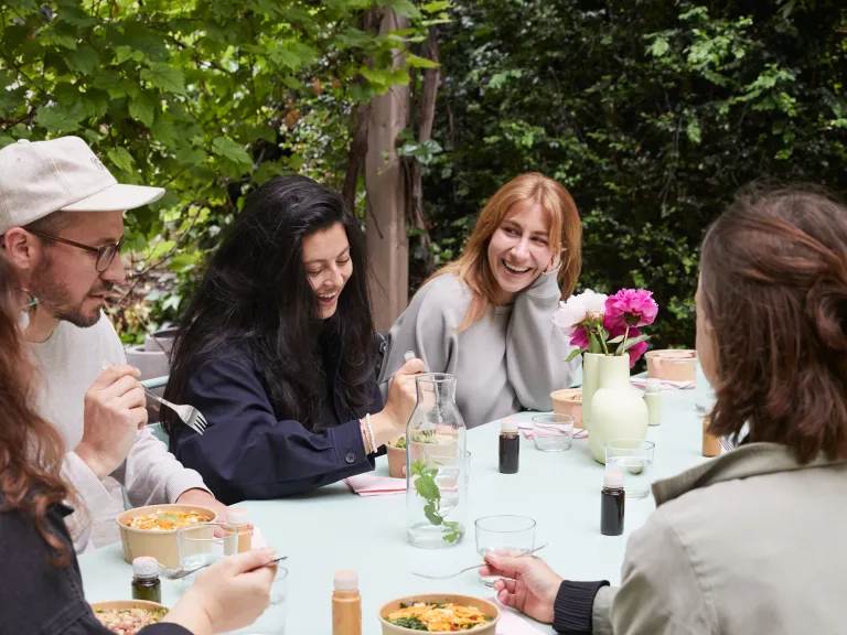 Eine Gruppe von Kolleg:innen geniesst gemeinsames Mittagessen an einem langen Tisch im Freien.