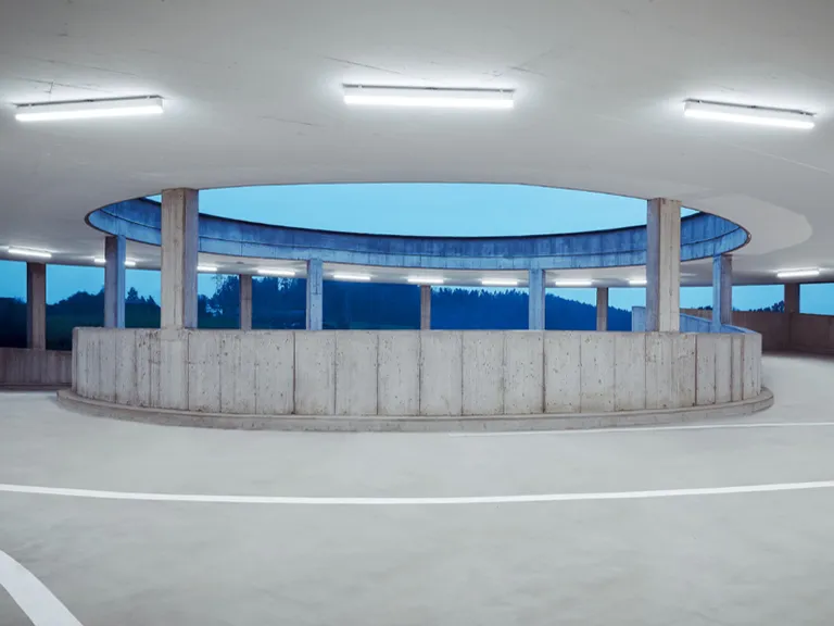 Ein breites, rundes Parkhaus aus Beton mit einem großen offenen Innenhof. Das minimalistische Design zeigt rohe Betonpfeiler und eine niedrige Schutzmauer, beleuchtet von horizontalen Leuchtstofflampen vor dem Dämmerungshimmel.