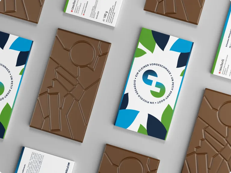 Draufsicht auf mehrere unverpackte Milchschokoladentafeln und ihre Verpackungen. Die Schokolade ist mit einem geometrischen, abstrakten Muster geprägt. Die Verpackungen zeigen das Holcim-Logo, umgeben von farbigen blau- und grünlichen, blattartigen Formen.