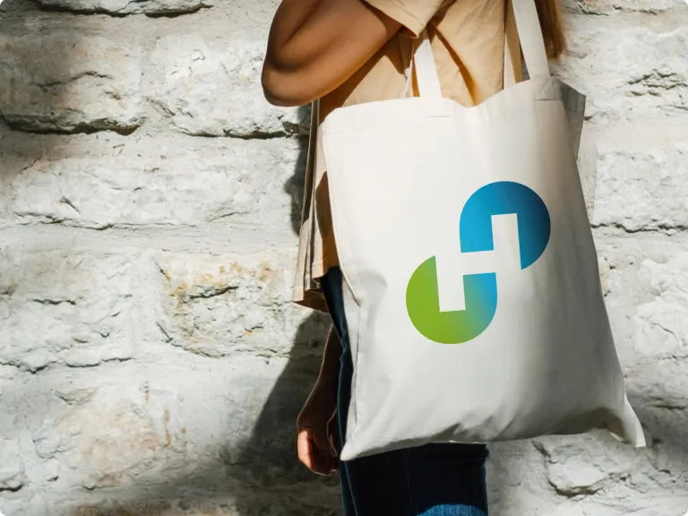 Eine Person trägt eine weiße Stofftasche über der Schulter und steht vor einer Steinwand. Auf der Tasche ist ein großes Holcim-Logo in einem Grün-Blau-Verlauf abgebildet.