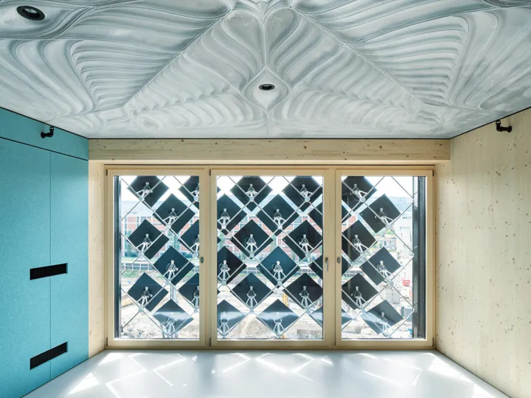 Ein Innenraum mit einer sehr detaillierten, gerippten Betondecke. Ein großes Fenster mit einer rautenförmigen Gitterstruktur aus Solarpaneelen gibt den Blick auf eine Baustelle frei.