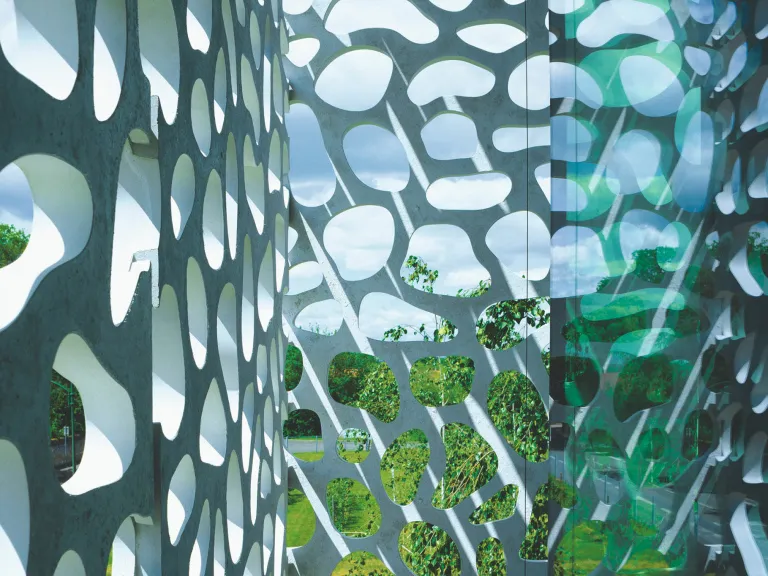 Nahaufnahme einer weißen Betonfassade mit organischen, kieselsteinförmigen Ausschnitten. Sonnenlicht fällt durch die Öffnungen.