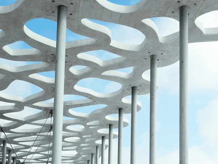 Aufnahme aus der Froschperspektive eines skulpturalen Betondachs, das von hohen, schlanken Pfeilern getragen wird. Das Dach weist ein poröses, netzartiges Muster mit großen, organischen Öffnungen auf und ist vor einem hellen, leicht bewölkten Himmel zu sehen.