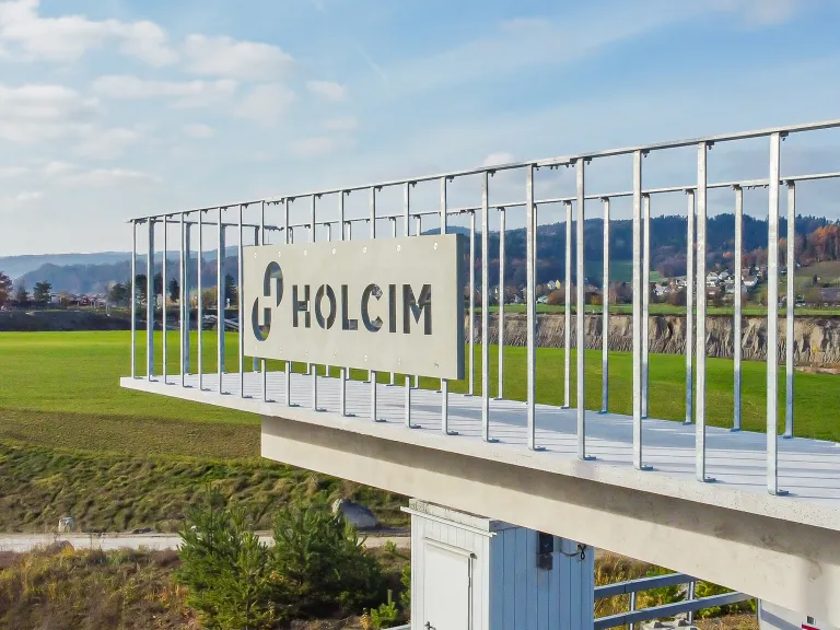 Eine Beton-Aussichtsplattform mit einem Metallgeländer, die auf ein grünes Tal und einen Steinbruch blickt. Ein graues Schild am Geländer zeigt das ‚HOLCIM‘-Logo als Ausstanzung im Material.