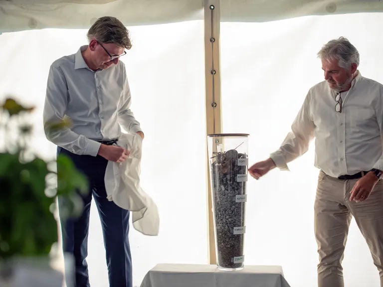  Zwei Männer in hellen Hemden stehen in einem weissen Zelt und flankieren einen hohen Glaszylinder, der Schichten verschiedener Mineral- und Metallfragmente enthält.